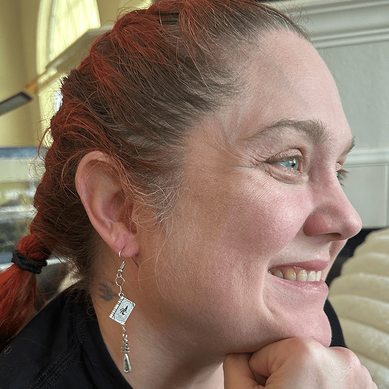 Model wearing Game Master Tome Gaming Earrings with silver book charm and hanging cudgel charm on chain.